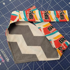 May include: A dog bandana featuring a gray and white chevron pattern on one side and a repeating pattern of the San Antonio Spurs logo on the other. The Spurs logo includes the text "SAN ANTONIO" and "SPURS".