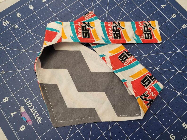 May include: A dog bandana featuring a gray and white chevron pattern on one side and a repeating pattern of the San Antonio Spurs logo on the other. The Spurs logo includes the text "SAN ANTONIO" and "SPURS".