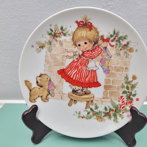 May include: A decorative plate featuring a young girl in a red and white striped dress hanging stockings by a brick fireplace. A kitten holds a stocking. The plate is decorated with Christmas greenery and candy canes.