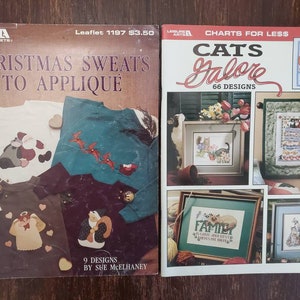 May include: Two craft books with cross stitch patterns. The book on the left is titled "Christmas Sweats to Applique" and features a white shirt with a red and white applique of a Santa Claus. The book on the right is titled "Cats Galore" and features a cross stitch pattern of a cat with the text "I love cats".