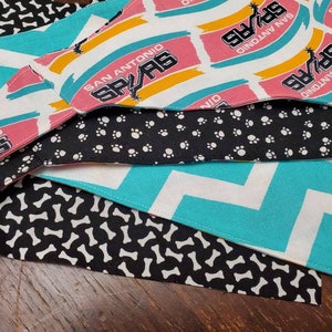 May include: A stack of dog bandanas in various patterns. One features the San Antonio Spurs logo in pink, black, and white. Other bandanas have patterns of white paw prints on black, white bones on black, and a turquoise chevron design.