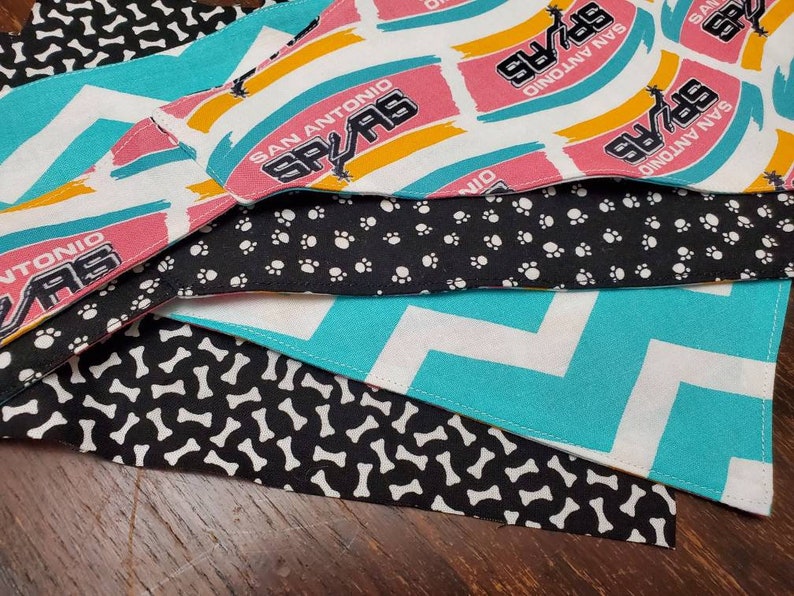 May include: A stack of dog bandanas in various patterns. One features the San Antonio Spurs logo in pink, black, and white. Other bandanas have patterns of white paw prints on black, white bones on black, and a turquoise chevron design.