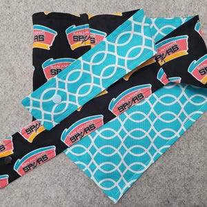 May include: A set of reusable fabric items featuring a black background with the San Antonio Spurs logo in red, yellow, and teal. Accents include teal fabric with a white geometric pattern. The set includes a variety of sizes and shapes.