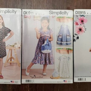 May include: Three Simplicity sewing pattern packages for girls' dresses. The patterns are for sizes 4-14. The first pattern is D0909 AA and features a pink, blue, and yellow dress. The second pattern is D0911 A and features a purple floral dress. The third pattern is D0915 A and features a pink floral dress.