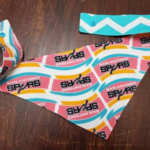 May include: A San Antonio Spurs themed bandana with a white background and repeating pink, yellow, and teal brushstroke design. The bandana features the team's logo and the words "SAN ANTONIO SPURS". The adjustable neck strap has a teal and white chevron pattern.