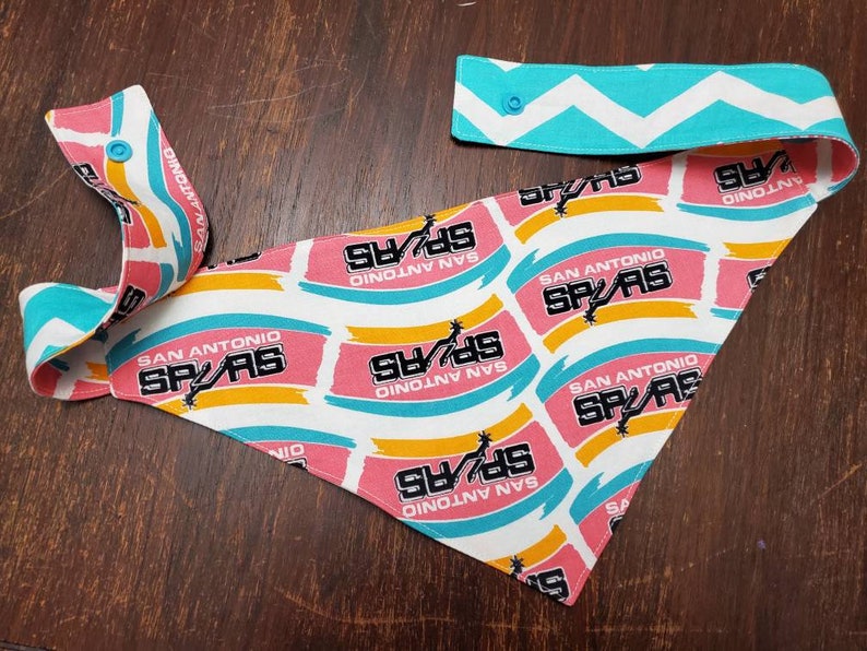 May include: A San Antonio Spurs themed bandana with a white background and repeating pink, yellow, and teal brushstroke design. The bandana features the team's logo and the words "SAN ANTONIO SPURS". The adjustable neck strap has a teal and white chevron pattern.