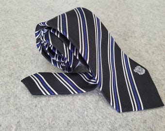 May include: A black tie with vertical stripes in white and blue. The tie is folded over itself, with a small emblem near the tip. The tie is lying on a gray surface.