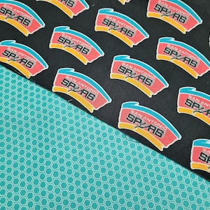 May include: Fabric featuring a repeating pattern of the San Antonio Spurs logo in red, yellow, and blue on a black background. A turquoise honeycomb pattern fabric is in the lower left corner. Ideal for crafting projects or apparel.