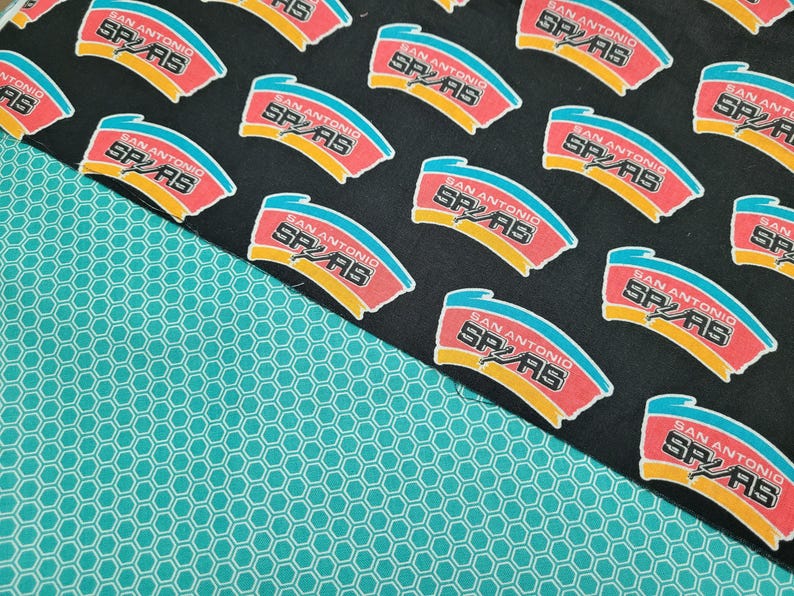 May include: Fabric featuring a repeating pattern of the San Antonio Spurs logo in red, yellow, and blue on a black background. A turquoise honeycomb pattern fabric is in the lower left corner. Ideal for crafting projects or apparel.