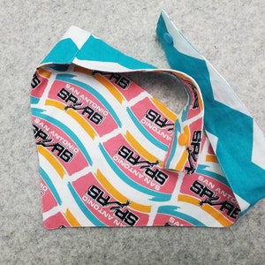 May include: A reversible pet bandana featuring a San Antonio Spurs team logo pattern on one side and a teal and white chevron pattern on the other. The bandana has a snap closure and is made of fabric.