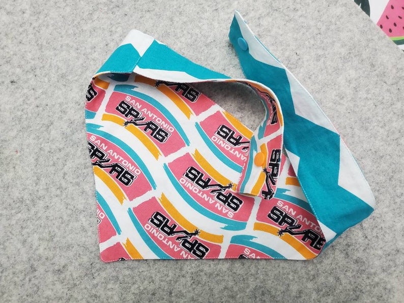 May include: A reversible pet bandana featuring a San Antonio Spurs team logo pattern on one side and a teal and white chevron pattern on the other. The bandana has a snap closure and is made of fabric.