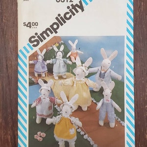 May include: Simplicity pattern number 6312 for making a set of seven bunny rabbit plush toys. The pattern includes instructions for making a variety of bunny rabbits in different outfits, including a blue dress, a yellow dress, a white dress, a blue and white striped outfit, and a pink and white striped outfit.