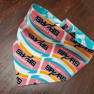 May include: A reversible bandana featuring the San Antonio Spurs logo and team name. The bandana has a white background with pink, teal, and yellow stripes. The reverse side is teal with a white zigzag pattern.