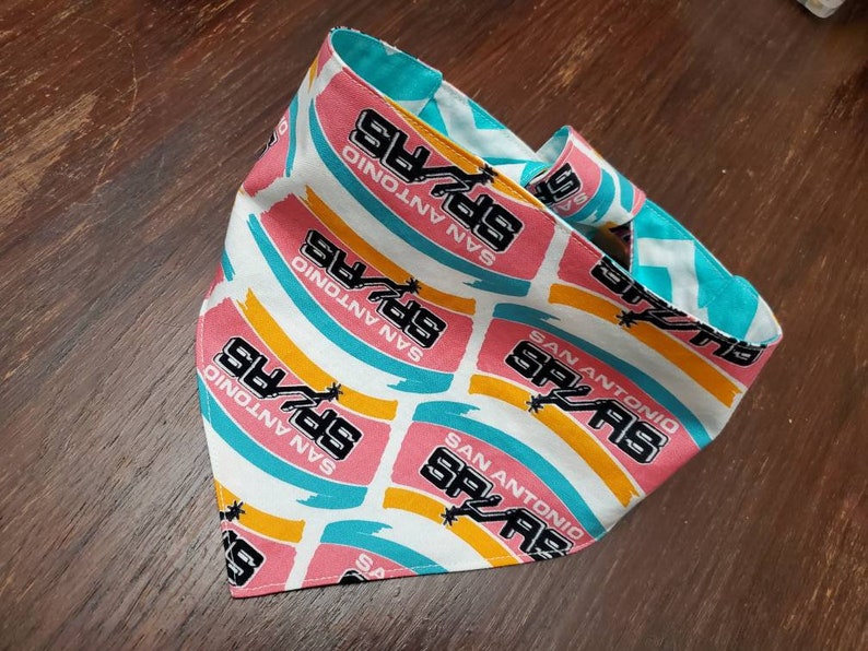 May include: A reversible bandana featuring the San Antonio Spurs logo and team name. The bandana has a white background with pink, teal, and yellow stripes. The reverse side is teal with a white zigzag pattern.