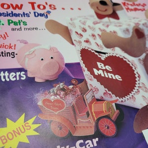 May include: A magazine cover with a pink piggy bank, a red heart with the text "Be Mine", and a red toy truck filled with candy hearts. The magazine cover has the text "27 Valentine Things To Do!" and "Candy-Car Cutout".