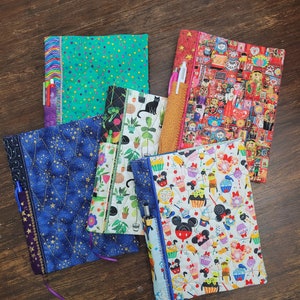 May include: Five different fabric-covered notebooks with purple ribbon bookmarks. The notebooks have a variety of patterns including stars, cupcakes, plants, and a colorful pattern with many small images.