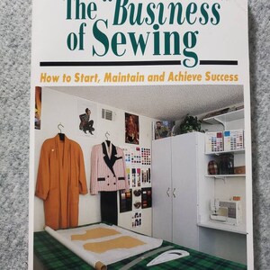 May include: A book titled "The Business of Sewing: How to Start, Maintain and Achieve Success" by Barbara Wright Sykes. The cover features a photo of a sewing room with a sewing machine, fabric swatches, and a garment on a dress form.