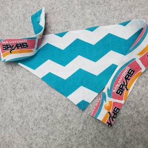 May include: A dog bandana featuring a teal and white chevron pattern. The ends display the San Antonio Spurs logo in pink, orange, and black. The bandana is secured with an orange button.