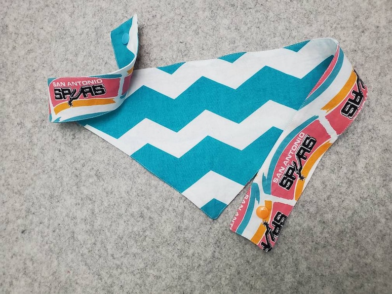 May include: A dog bandana featuring a teal and white chevron pattern. The ends display the San Antonio Spurs logo in pink, orange, and black. The bandana is secured with an orange button.