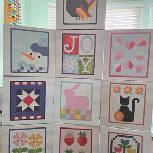 May include: A collection of ten square cards featuring seasonal quilt-style patterns, including a turkey, a snowman, the word JOY with a snowflake, hearts, a star, a rabbit, a black cat, flowers, strawberries, and a basket of tulips.