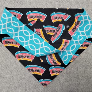 May include: A reversible dog bandana featuring the San Antonio Spurs logo on a black background. The reverse side has a turquoise and white geometric pattern. The bandana is folded and ready to wear.