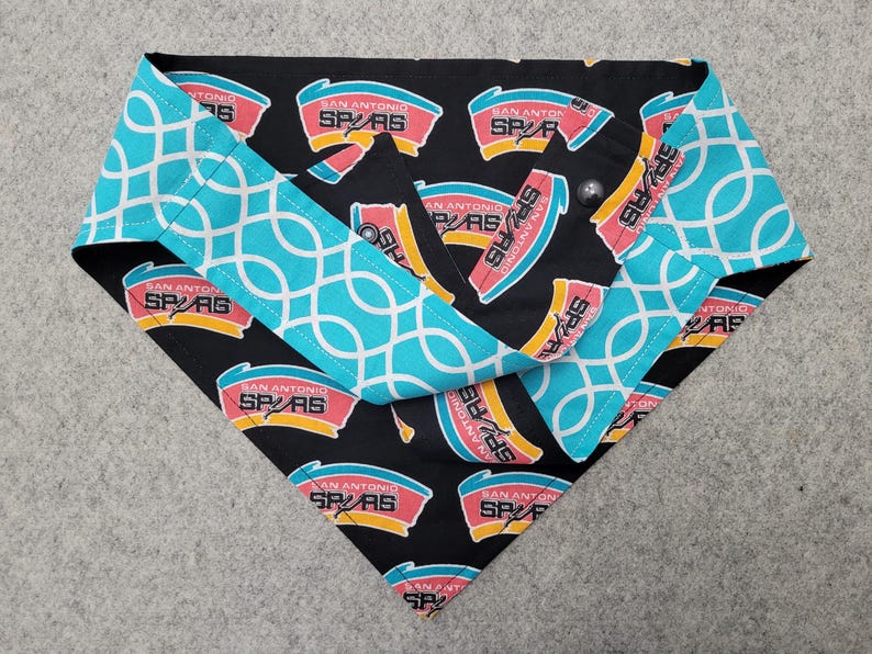 May include: A reversible dog bandana featuring the San Antonio Spurs logo on a black background. The reverse side has a turquoise and white geometric pattern. The bandana is folded and ready to wear.
