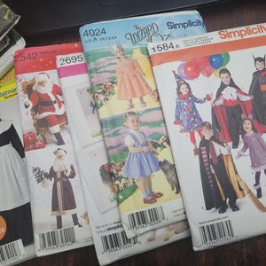 May include: A collection of Simplicity sewing patterns featuring costume designs. The patterns include images of children in various costumes, including Santa Claus, a witch, and characters from "The Wizard of Oz". The patterns are in various sizes.