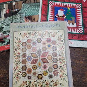 May include: Three quilting books with colorful covers. The book on the left is titled "Holiday Quilts" and features a green and white quilt. The book in the middle is titled "Handfuls of Scraps" and features a quilt with a floral border and a patchwork center. The book on the right is titled "More Joy to the World" and features a red and white quilt with a Santa Claus and a Christmas tree.