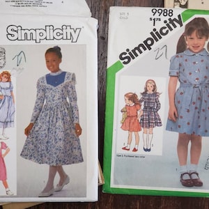 May include: Two vintage Simplicity sewing pattern envelopes. One is size 7, featuring a girl in a blue floral dress. The other is size 5, with a girl in a blue dress with red heart shapes. Both patterns are for children's clothing.