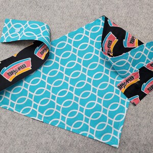 May include: A handmade pet bandana featuring a turquoise and white geometric pattern, with a black and white trim displaying the San Antonio Spurs logo. The bandana is folded and laid out on a gray surface.