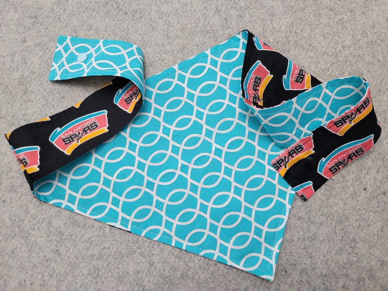 May include: A handmade pet bandana featuring a turquoise and white geometric pattern, with a black and white trim displaying the San Antonio Spurs logo. The bandana is folded and laid out on a gray surface.