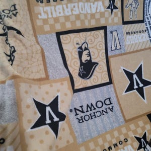 May include: A tan and cream-colored fabric with various square and rectangular patches. The patches feature the text "VANDERBILT COMMODORES" and "ANCHOR DOWN" along with star and hand graphics. The letter "V" is also prominently displayed.