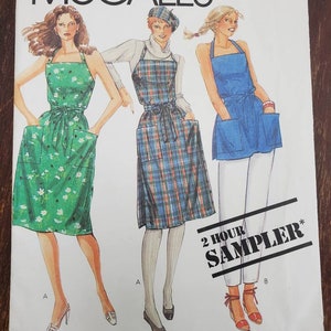 May include: A vintage McCall's sewing pattern featuring three women's clothing designs. The first design is a green wrap dress with white floral print. The second design is a plaid apron with a black belt. The third design is a blue apron with white pockets. The pattern is titled "2 Hour Sampler*" and includes the text "Misses' Wrap Dress and Top".