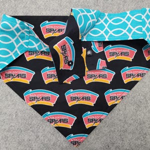 May include: A dog bandana featuring a black background with the San Antonio Spurs logo in red, yellow, and teal. The bandana has teal and white geometric patterned accents on the collar.