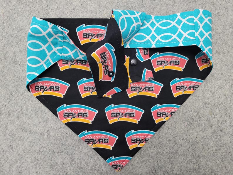 May include: A dog bandana featuring a black background with the San Antonio Spurs logo in red, yellow, and teal. The bandana has teal and white geometric patterned accents on the collar.