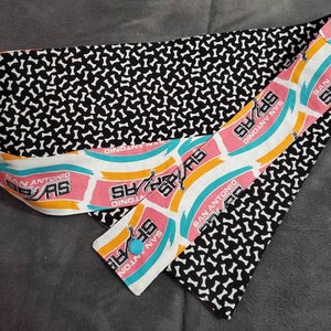 May include: A fabric headband featuring a black and white bone pattern and a section with the San Antonio Spurs logo in pink, teal, and yellow. The headband has a button closure and is designed for comfort and style.