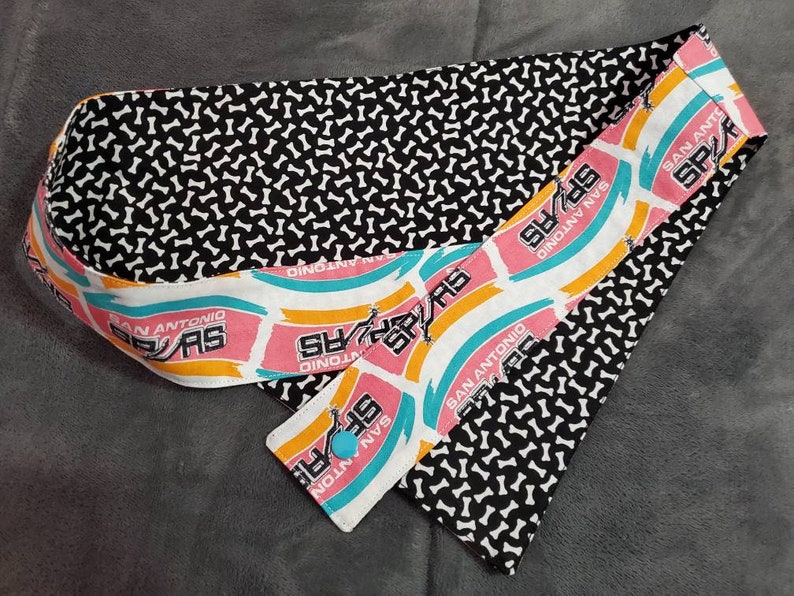 May include: A fabric headband featuring a black and white bone pattern and a section with the San Antonio Spurs logo in pink, teal, and yellow. The headband has a button closure and is designed for comfort and style.