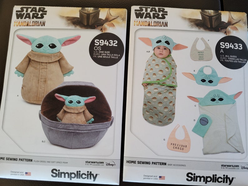 May include: Two Simplicity sewing patterns for Star Wars: The Mandalorian. The left pattern is for a plush Grogu toy and a soft space pram. The right pattern is for a baby hooded towel, a bib, and a sleeping bag. The patterns are numbered S9432 and S9433.