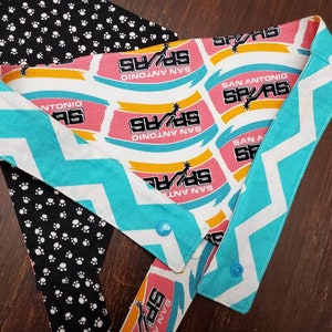 May include: A dog bandana featuring the San Antonio Spurs logo in pink, yellow, and white. The bandana has a turquoise and white chevron pattern on the edges and a black and white paw print pattern on the back.