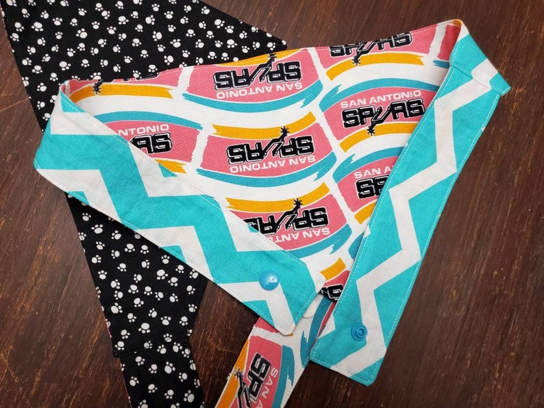 May include: A dog bandana featuring the San Antonio Spurs logo in pink, yellow, and white. The bandana has a turquoise and white chevron pattern on the edges and a black and white paw print pattern on the back.
