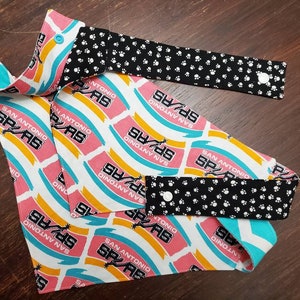 May include: A reversible pet bandana and matching face mask featuring a repeating pattern of the San Antonio Spurs logo in pink, white, and teal. The reverse side is black with white paw prints. Both pieces have white snap closures.