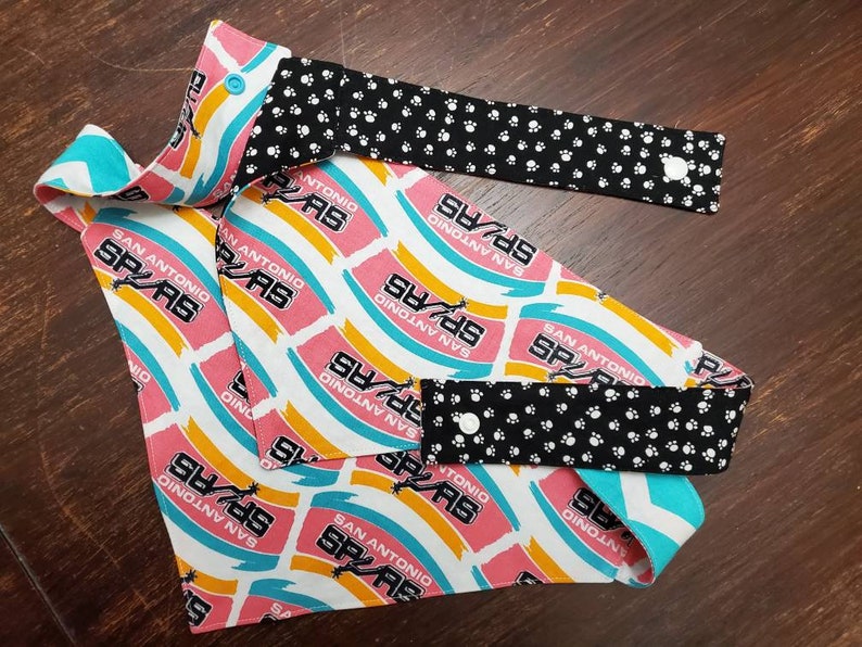 May include: A reversible pet bandana and matching face mask featuring a repeating pattern of the San Antonio Spurs logo in pink, white, and teal. The reverse side is black with white paw prints. Both pieces have white snap closures.