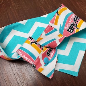 May include: A reversible fabric item with a turquoise and white chevron pattern on one side and a colorful San Antonio Spurs logo print on the other. The item has a blue button closure.