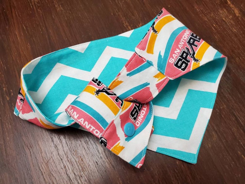 May include: A reversible fabric item with a turquoise and white chevron pattern on one side and a colorful San Antonio Spurs logo print on the other. The item has a blue button closure.