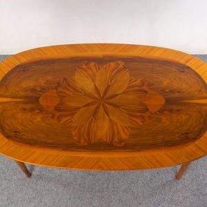 May include: Oval wooden coffee table with a floral design inlay. The table features a light brown border and a darker brown center with a flower-like pattern. The table is supported by four wooden legs.