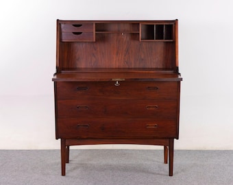 May include: A dark brown wooden secretary desk featuring a drop-down writing surface, three lower drawers with horizontal handles, and an upper hutch section containing two small drawers, an open shelf, and three vertical storage compartments.