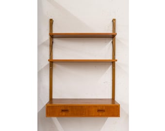 Puede incluir: Una estantería de madera montada en la pared que cuenta con dos estantes horizontales y una sección de escritorio inferior con dos cajones integrados. La unidad se apoya en dos rieles verticales de madera fijados a la pared, mostrando una veta de madera marrón cálida.