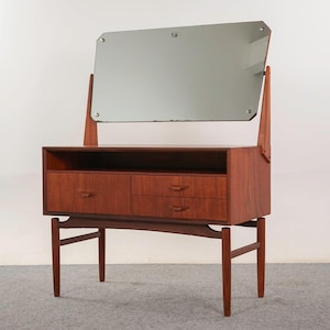 Peut inclure: Une coiffeuse en bois de style moderne du milieu du siècle avec un miroir rectangulaire. La coiffeuse a deux tiroirs et une étagère. Le miroir est fixé avec des supports en bois. Le meuble est en bois aux tons chauds et a un design minimaliste.