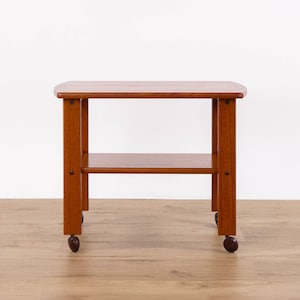 Peut inclure: Une table d'appoint rectangulaire en bois avec une finition brun moyen lisse. La table présente une surface supérieure plate, une étagère inférieure pour le rangement et quatre pieds verticaux. Chaque pied est muni d'une petite roulette.