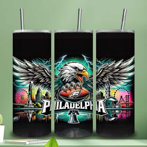 May include: Black insulated tumbler featuring a vibrant graphic of a bald eagle with outstretched wings, clutching a football. The design includes the city skyline, the word "PHILADELPHIA," and a football helmet. The tumbler has a silver straw.